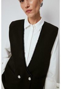 Black vest with a deep V-neck, trimmed in round black beads and secured with silver buttons, layered over a white collared shirt.