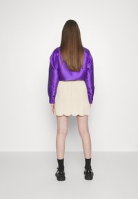 Veste bomber en satin violet avec manches bouffantes, associée à une jupe beige à ourlet festonné et des chaussettes noires jusqu'à la cheville portées avec des chaussures noires épaisses.