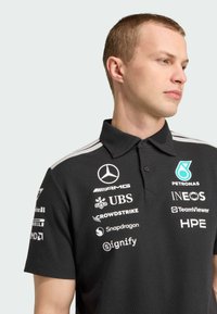 Polo noir à manches courtes, avec un col à trois boutons et plusieurs logos de sponsors en blanc et teal. Des rayures grises ornent les épaules.