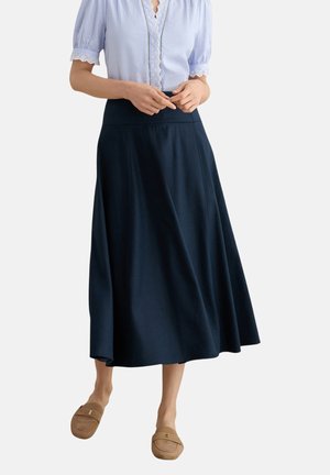 Frau trägt einen marineblauen Midirock, eine hellblaue Bluse mit kurzen Ärmeln und Spitzenbesatz sowie beige Slipper, steht vor einem einfarbigen Hintergrund.