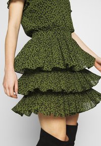 Vestido verde con estampado floral y volantes, que presenta una cintura ajustada y una textura transparente. El diseño incluye mangas cortas y una silueta en capas.