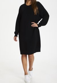 Abito nero a costine con maniche lunghe, scollo rotondo e vestibilità rilassata, che raggiunge la metà della coscia. Abbinato a sneakers bianche.