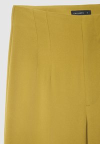 Pantalon taille haute jaune, cintré, avec une texture douce, présentant des plis marqués à l'avant et une étiquette discrète sur la ceinture.