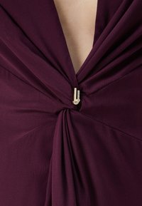 Šaty s hlbokým dekoltom v tmavo červenej farbe, so zvlnenou látkou, centrálnym zvratom a dekoratívnym kovovým sponou so trblietavými akcentmi.