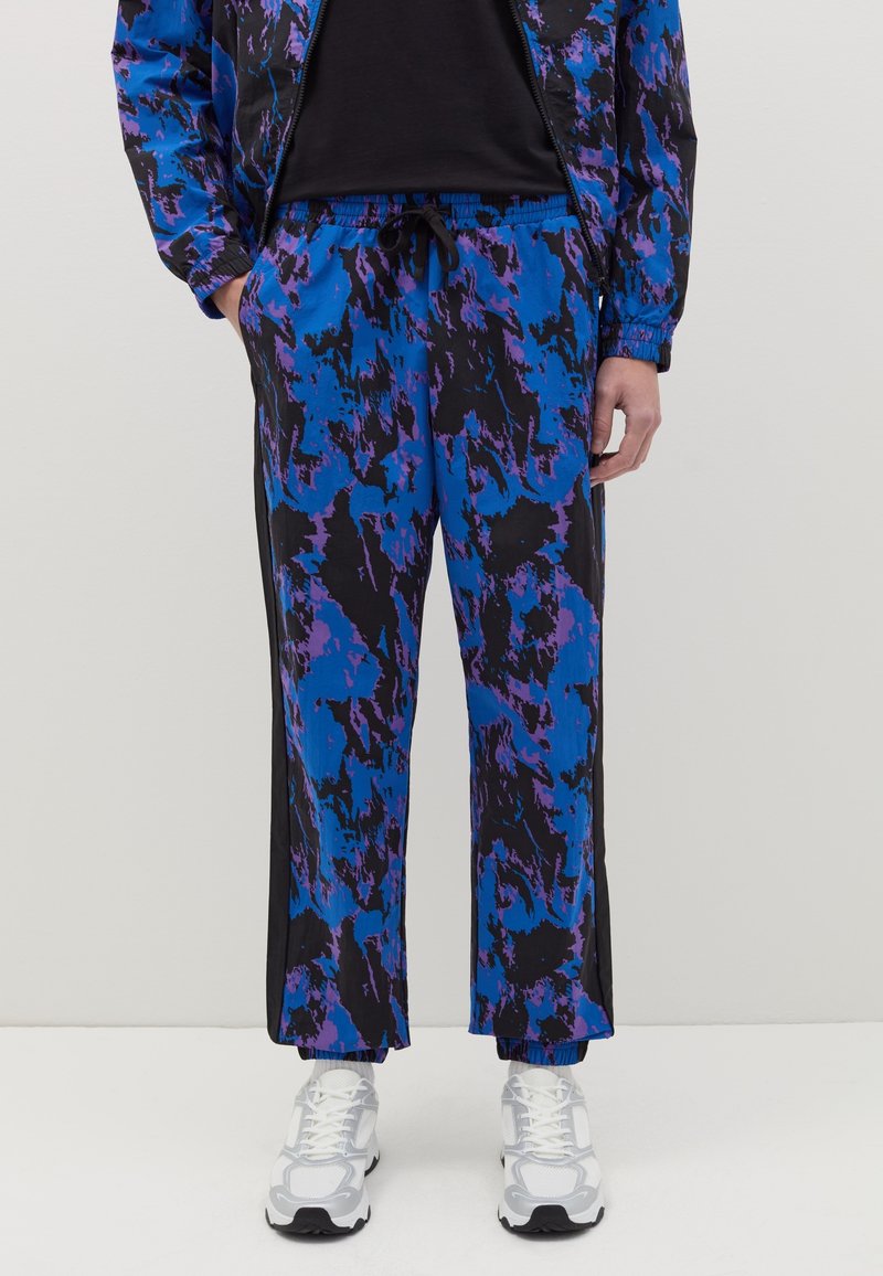 Pantalons de survêtement avec un motif abstrait bleu et noir, taille élastique et rayures sur les côtés. Fabriqués en tissu léger ; portés avec des baskets blanches.