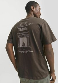 Brown cotton T-shirt with short sleeves, featuring a fireplace graphic and text on the back. Relaxed fit with a soft texture.