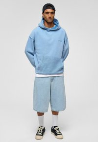 Lichtblauwe hoodie met een kangoeroe-pocket, gecombineerd met lichtdenim shorts. Het model draagt zwarte schoenen met crèmekleurige accenten en witte sokken.