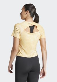 Top atletico a maniche corte giallo tie-dye con design aperto sul retro, che mostra un reggiseno sportivo nero sottostante. Indossato con leggings neri aderenti.