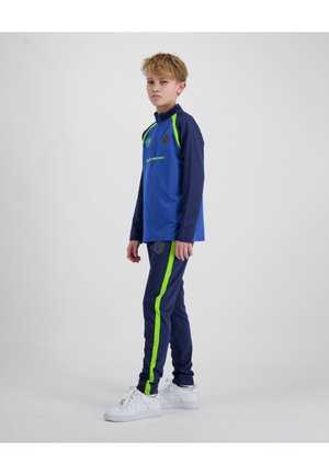 Jongen staand opzij, gekleed in een blauw en marineblauw trainingspak met neon groene strepen en witte sneakers, tegen een effen lichtgrijze achtergrond.