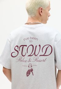 Grå oversized t-shirt med maroon tekst på ryggen: "Jeg har været på STOWD Relax & Resort for Krop & Sind" med grafiske detaljer.