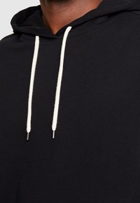 Sudadera con capucha negra que cuenta con una capucha amplia y cordones ajustables en color crema con extremos metálicos. Textura de tela suave, diseño minimalista.