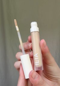 Il correttore ILIA in un tubo trasparente con un tappo bianco, dotato di un applicatore a forma di piede di cervo. La formula appare di un beige chiaro con una texture liscia.
