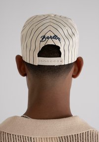 Vue arrière d'une personne portant une casquette beige à rayures avec "Berlin" brodé et un t-shirt en tricot texturé beige sur un fond uni.