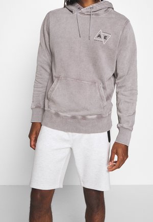 Mann trägt einen ausgebleichten grauen Hoodie mit Fronttasche und AE-Logo, kombiniert mit weißen Shorts bis zum Knie.