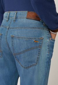 Jeans in denim blu con una vita scura, caratterizzati da un leggero sbiadimento, cuciture arancioni e un piccolo logo di un bassotto ricamato sulla tasca posteriore.