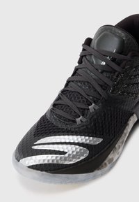 Baskets noires de sport avec une tige en maille texturée, des lacets plats et une semelle en caoutchouc transparent. Présente un accent argenté brillant sur le côté.