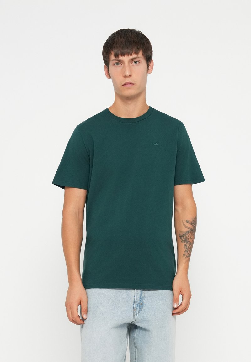 Giovane uomo con capelli corti scuri, che indossa una maglietta verde scuro e jeans azzurri chiari, in piedi su uno sfondo bianco uniforme.