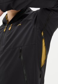 Zwarte jas met gele binnenvoering, voorzien van een staande kraag, ritszakken en een gladde, gestructureerde stof. Accenten omvatten ritsen en merking.