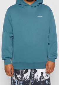 Hombre con sudadera teal de Calvin Klein y pantalones cortos de patrón abstracto en negro, blanco y azul, de pie con las manos relajadas a los lados.