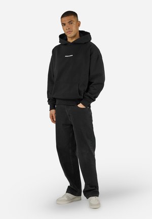 Νεαρός άνδρας που στέκεται με το ένα χέρι στην τσέπη, φοράει μαύρο φούτερ Pegador, χαλαρά μαύρα τζιν και λευκά αθλητικά παπούτσια σε λευκό φόντο.