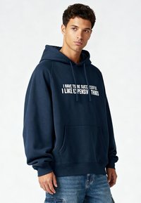 Sweat à capuche bleu marine en coton, avec une poche avant, une capuche à cordon de serrage et un texte imprimé en blanc sur la poitrine. Coupe ample et texture douce.