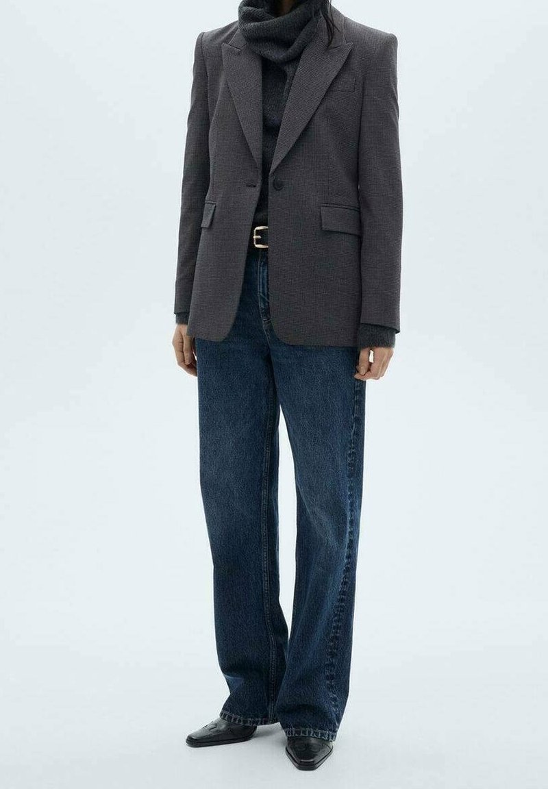 Personne portant un blazer gris foncé, un pull à col roulé noir, un jean large bleu avec ceinture noire et des chaussures pointues noires sur fond blanc.