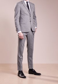 Traje gris a medida con un acabado texturizado, chaqueta de un solo botón, solapas y pantalones de corte slim. Combinado con zapatos de vestir de cuero negro.