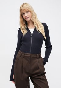 Maglione blu navy a costine con zip, maniche lunghe e dettagli sulle spalle, abbinato a pantaloni marroni a vita alta. Tessuto liscio, design aderente.