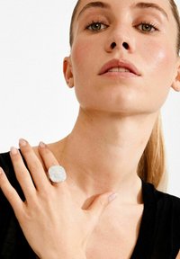 Sõrmus, millel on tekstureeritud, hõbedase värvusega pind ja ruudukujuline kuju. Kandmisel on see käel, millel on loomulikud küüned, neutraalse taustaga.