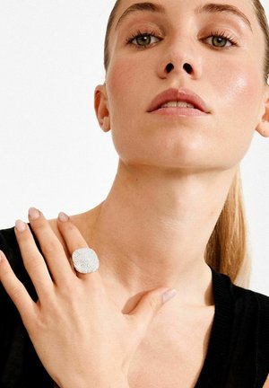 Sõrmus, millel on tekstureeritud, hõbedase värvusega pind ja ruudukujuline kuju. Kandmisel on see käel, millel on loomulikud küüned, neutraalse taustaga.