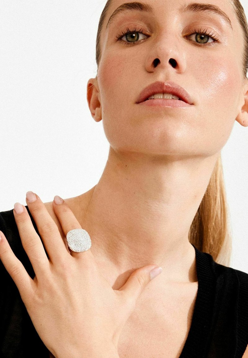 Sõrmus, millel on tekstureeritud, hõbedase värvusega pind ja ruudukujuline kuju. Kandmisel on see käel, millel on loomulikud küüned, neutraalse taustaga.