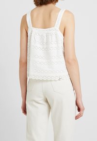 Top sin mangas de algodón blanco con dobladillo festoneado y bordado de ojales con rayas horizontales. Se lleva con pantalones de color claro.