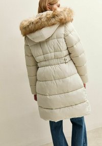 Mujer vestida con un abrigo largo acolchado beige, con capucha con borde de piel y cintura ajustada, combinado con jeans azules, de pie frente a una pared lisa.