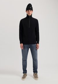 Pullover nero a coste con zip e collo alto, abbinato a jeans blu e sneakers grigie. Presenta un design aderente e una texture a maglia.