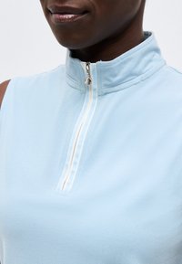 Κοντινό πλάνο ενός ατόμου που φοράει αμάνικο ανοιχτό γαλάζιο τοπ με φερμουάρ στον ψηλό γιακά και λευκές ρίγες κατά μήκος του φερμουάρ.