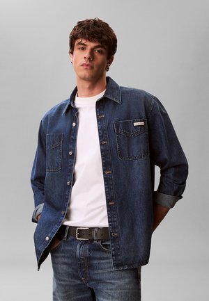 Młody mężczyzna ubrany w ciemnoniebieską kurtkę jeansową na białej koszulce i niebieskich dżinsach z czarnym paskiem, stojący na jednolitym szarym tle.