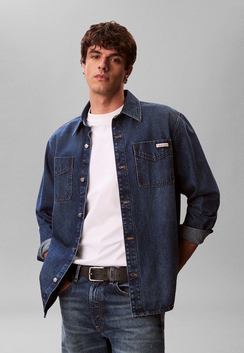 Ung mand iført en mørkeblå denimjakke over en hvid t-shirt og blå jeans med sort bælte, stående foran en ensfarvet grå baggrund.