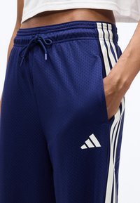 Κοντινό πλάνο ενός ατόμου που φοράει σορτς Adidas από δίχτυ σε ναυτικό μπλε, με άσπρες ρίγες και λογότυπο, το χέρι χαλαρά στην πλαϊνή τσέπη.