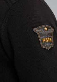 Czarny sweter z dzianiny, charakteryzujący się teksturowanym materiałem oraz naszywką z haftem, na której widnieje napis "PME", gwiazda oraz wzory skrzydeł w kolorze szarym i pomarańczowym.