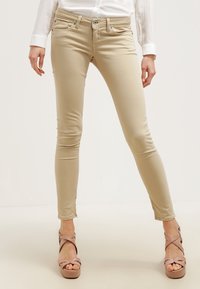 Žena oblečená v úzkých béžových kalhotách, bílé blůze a růžových platformových sandálech stojící na světlém pozadí.