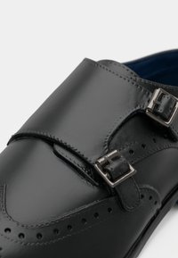 Chaussure habillée en cuir noir avec double brides de type moine et boucles argentées, présentant des détails brogue sur le bout et les côtés.