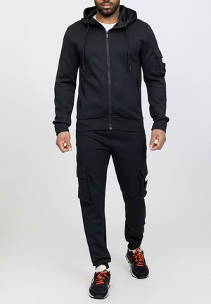 Ensemble de sport noir comprenant une veste à capuche zippée et un pantalon cargo avec poches. Fabriqué en tissu texturé, associé à des baskets noires et orange.