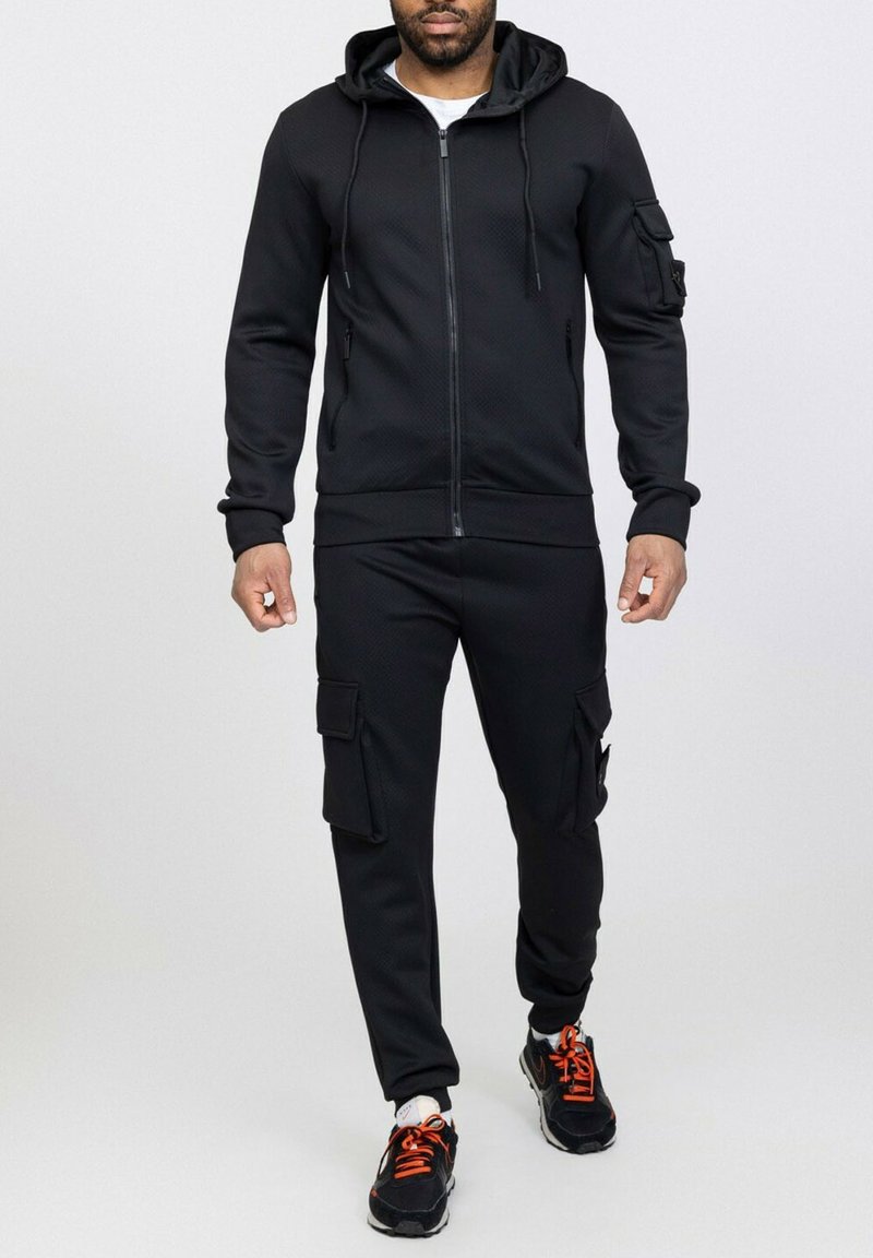 Ensemble de sport noir comprenant une veste à capuche zippée et un pantalon cargo avec poches. Fabriqué en tissu texturé, associé à des baskets noires et orange.