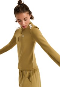 Lange mouwen sporttop in kaki met gestructureerde stof, kwart-rits nek en witte schouderaccents. Coördinerende ontspannen broek zichtbaar.