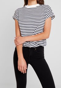 T-shirt a righe bianco e blu navy, maniche corte con risvolti, scollatura a girocollo. Abbinato a jeans neri, che evidenziano una silhouette aderente.