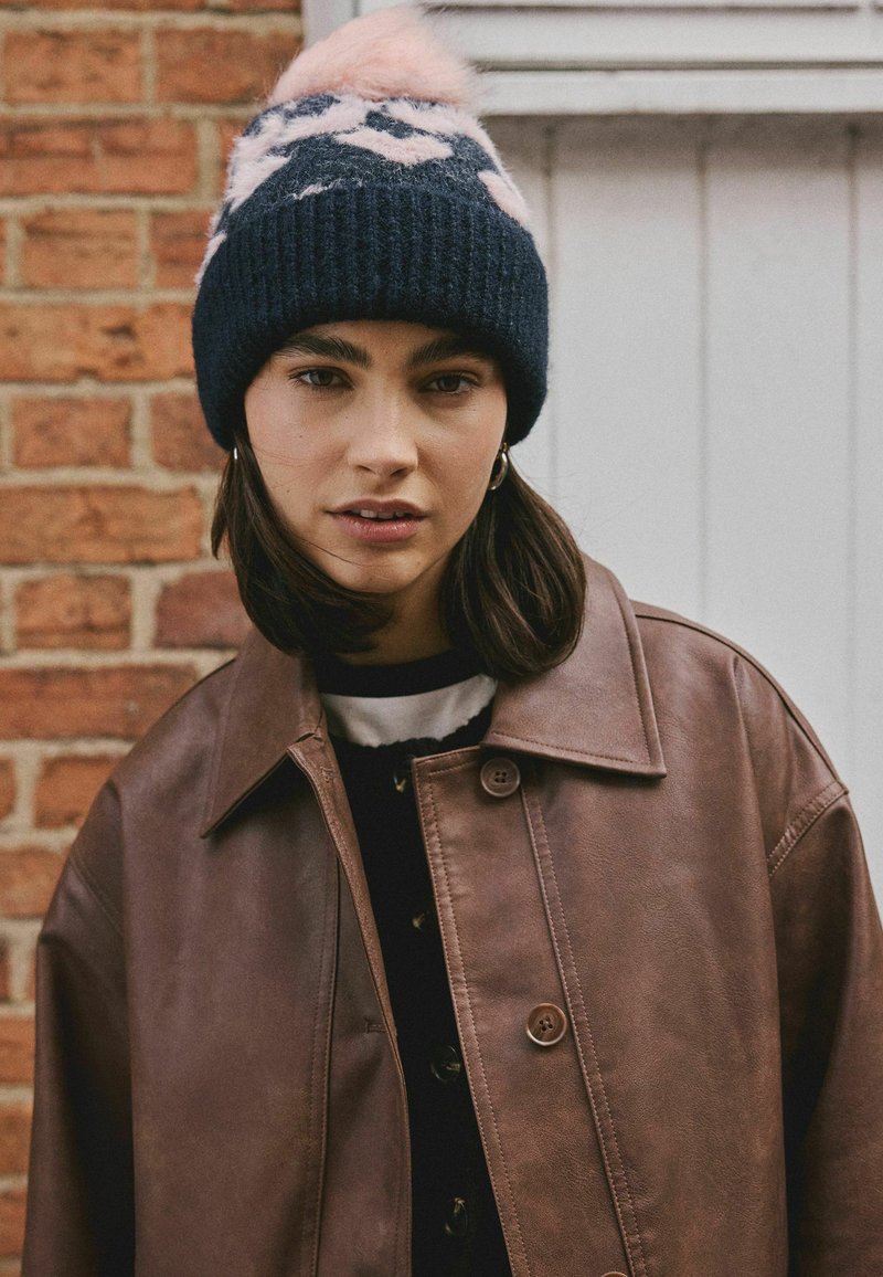 Jauna sieviete brūnā ādas jakā un tumši zila adīta cepurē ar rozā rakstiem stāv pretī ķieģeļu sienai un baltām durvīm.