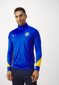 Giacca Nike a zip blu e gialla con colletto alto, logo sul petto, strisce laterali e polsini aderenti; texture del tessuto liscia.