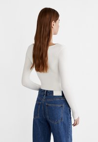 Kvinde med langt brunt hår iført en tætsiddende hvid langærmet top og højtaljede blå jeans, set fra bagenden mod en hvid baggrund.