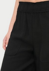 Gros plan d'une personne portant un pantalon noir taille haute avec une ceinture élastique et de grandes poches avant, main reposant près de la hanche.