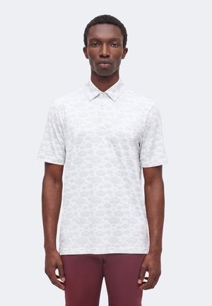 Homme aux cheveux courts et bouclés portant un polo blanc à manches courtes avec un motif subtil de couronne et un pantalon bordeaux sur fond uni.